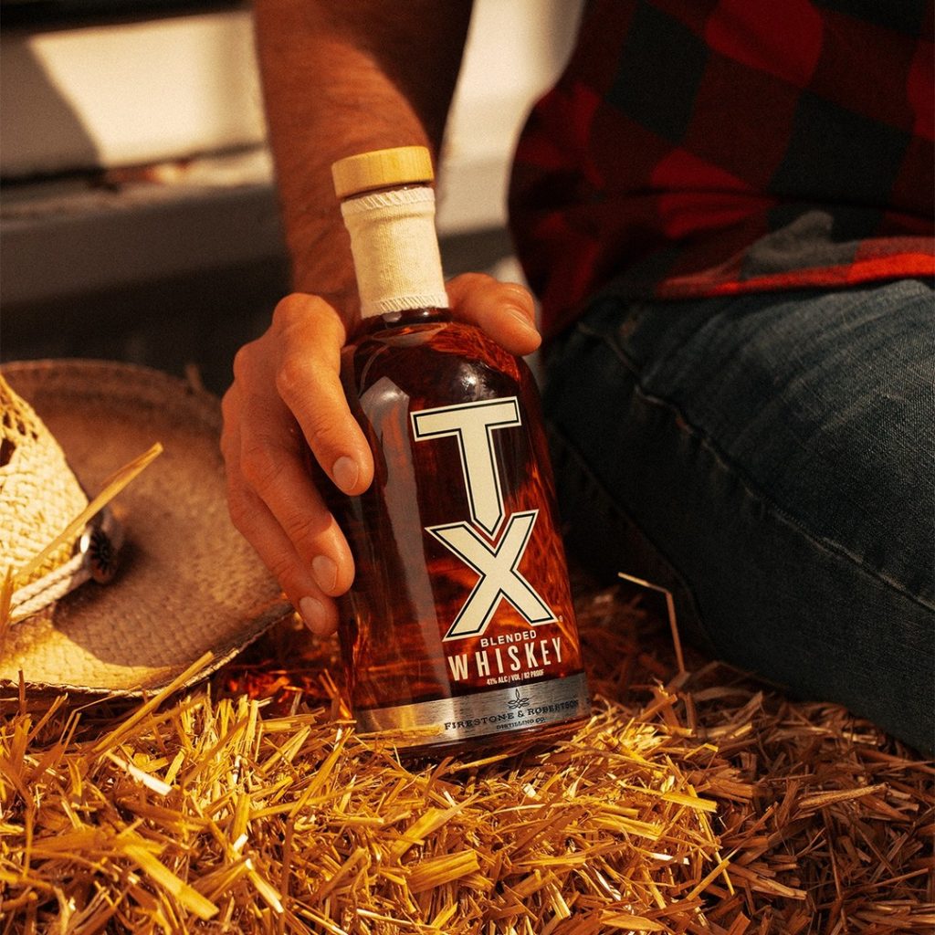 Close up shot of a man's hand holding a bottle of TX Whiskey with a distinctive canvas neckwrap. He sits atop a bale of hay, and in the frame you can also see his denim jeans, red and black plaid shirt, and a cowboy hat set down on the hay. 