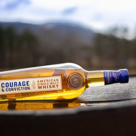 Bottle of Virginia Single Malt Whisky laying on it's side on top of a whisky barrel and the Virginia mountains blurred in the background. 