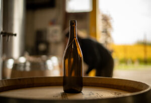 An empty amber Revino refillable bottle is seen sitting on top of a wine barrel with an unfocused background wherein a worker is seen looking at other wine barrels.